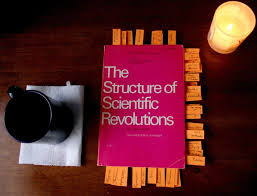 A copy of Thomas Kuhn's The Structure of Scientific Revolutions resting on a dark wooden desk beside a candle and a coffee cup